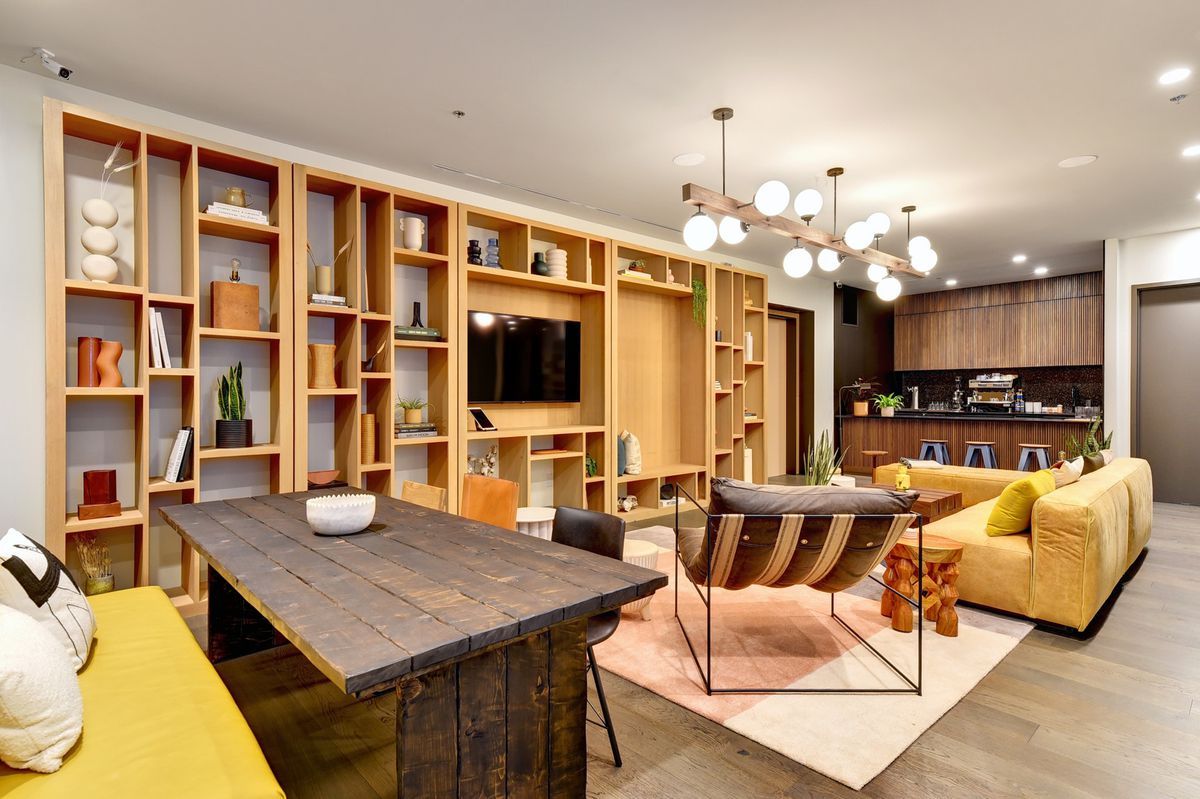A room set up as an apartment unit. It has a small kitchen with a breakfast bar and upper cabinets at one end. A giant shelving unit with various decorative items and a TV on it takes up one long wall. A couch and other furniture are arranged to face it. A bench-like seat and rustic dining table are on the close end. There’s a modern pendant light with globes sticking out of a central section hanging from the ceiling.