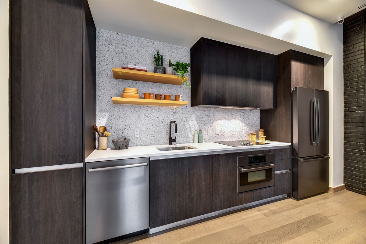 A small kitchen with upper and lower cabinets, appliances, sink and a pantry line up on one wall. There is a tile backsplash from countertop to ceiling and two open shelves on the wall. The floor is wood. Part of a brick wall makes a corner on the right side of the space.