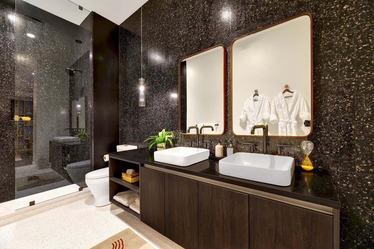 A large bathroom with floor-to ceiling wall covering and shower enclosure, a cabinet with rectangular double sinks, a mod toilet with a cylindrical glass pendant light above it, and a tall shower with a tall glass door.